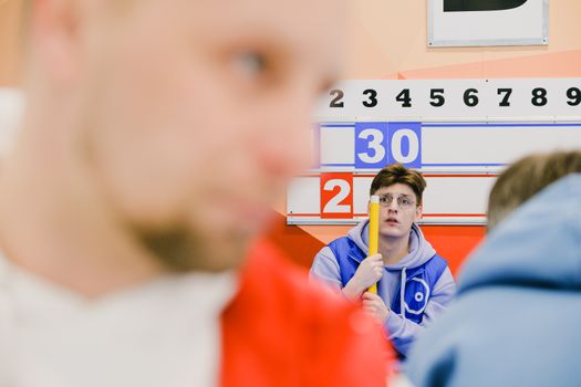 Blurred sportsman near male player with worrier face and broom in hands sitting near score board during sportive curling game