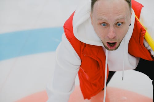 Amazed man in sportswear with opened mouth and broom playing curling on ice sheet with circular target area during game in playing area