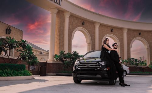 Free Stylish couple poses on a black car in an elegant setting under a dramatic sky. Stock Photo
