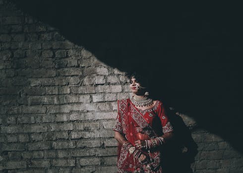 Elegant Indian bride in a red sari with ornate jewelry, leaning against a brick wall, exuding traditional charm.