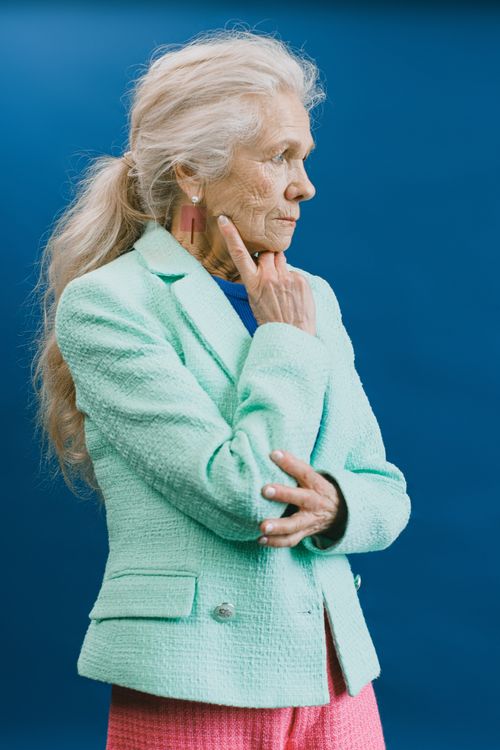 Free Side view of elderly gray haired lady with ponytail wearing green jacket touching chin and looking away standing against blue background Stock Photo