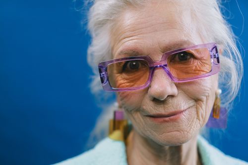 Free Headshot of kind elderly gray haired lady wearing eyeglasses and earrings looking at camera against blue background Stock Photo