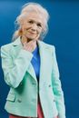 Senior female model with gray hair wearing green jacket and blue shirt standing near blue wall and touching chin while looking at camera