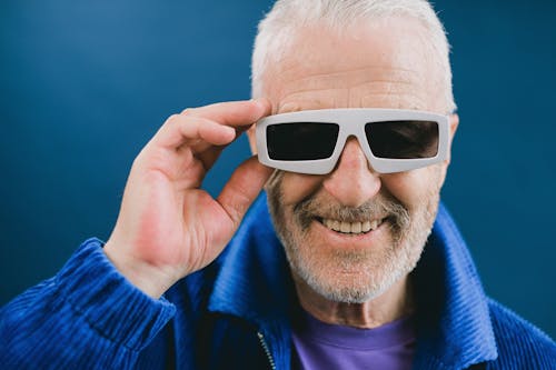 Portrait of smiling elderly unshaven male with gray hair wearing blue jacket and sunglasses looking at camera against blue background