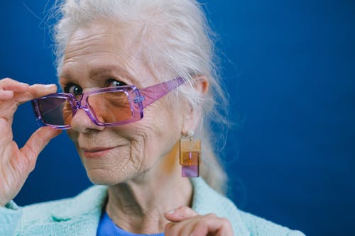 Retrato gratis de una mujer mayor elegante con cabello gris, usando gafas de sol moradas y aretes, sonriendo y mirando a la cámara contra un fondo azul - Foto de stock