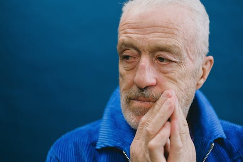 Portrait of elderly bearded gray haired male in blue jacket standing against dark blue background putting hands together in front of face and looking away
