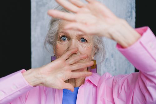 Free Close-up of an elderly woman with blue eyes in a pink shirt, hands in front of her face. Stock Photo