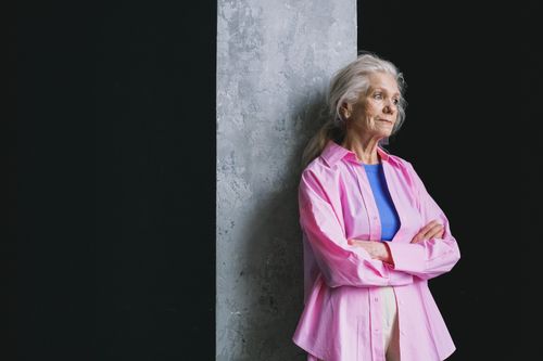 Free Senior woman with grey hair leaning against a wall, wearing a pink shirt, arms crossed, looking thoughtful. Stock Photo