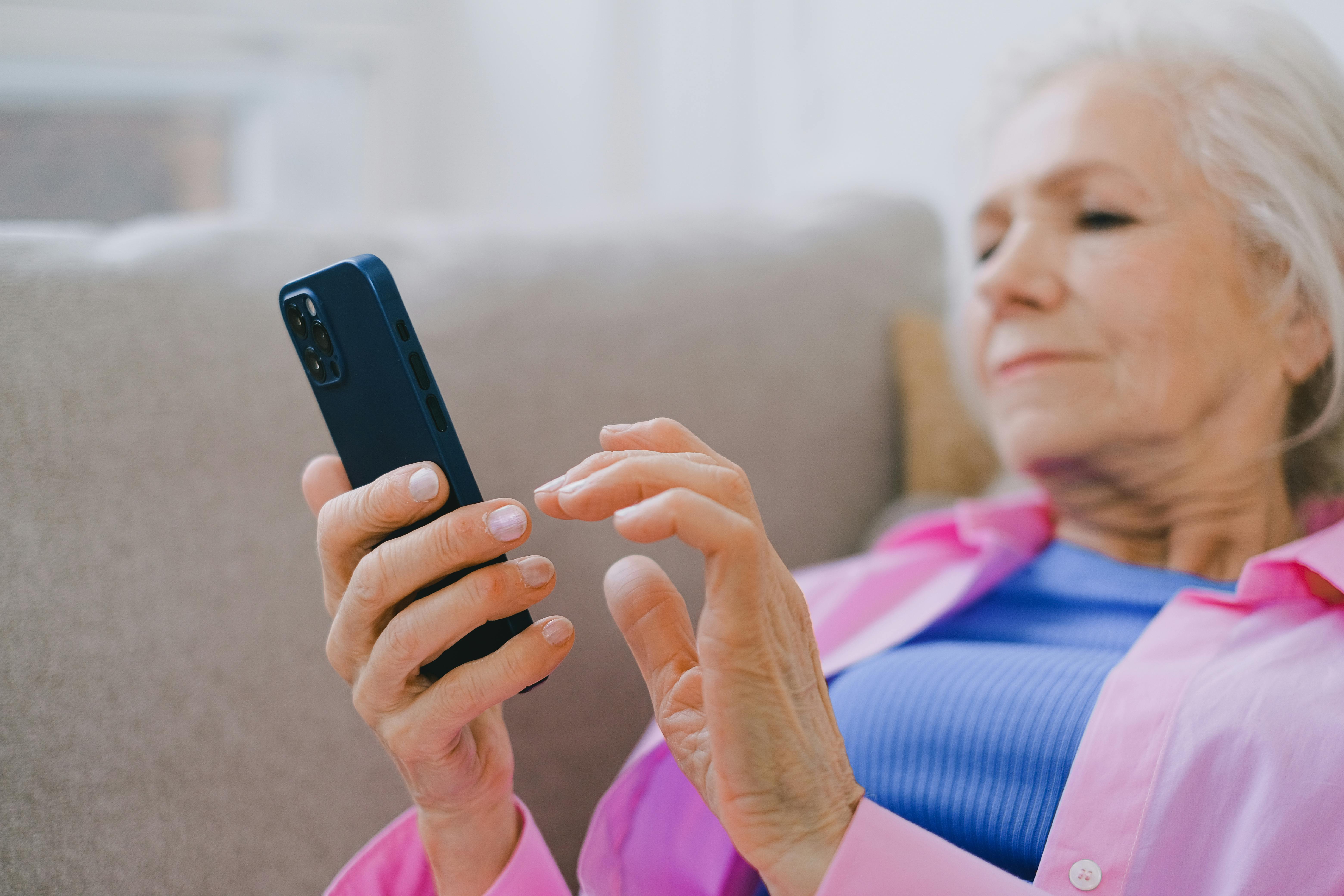 Elderly woman in pink shirt using smartphone on a couch at home, engaged in technology.