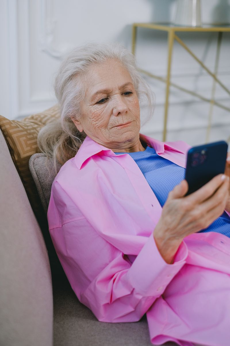 Elderly woman in pink using smartphone on couch, comfortably enjoying her time.