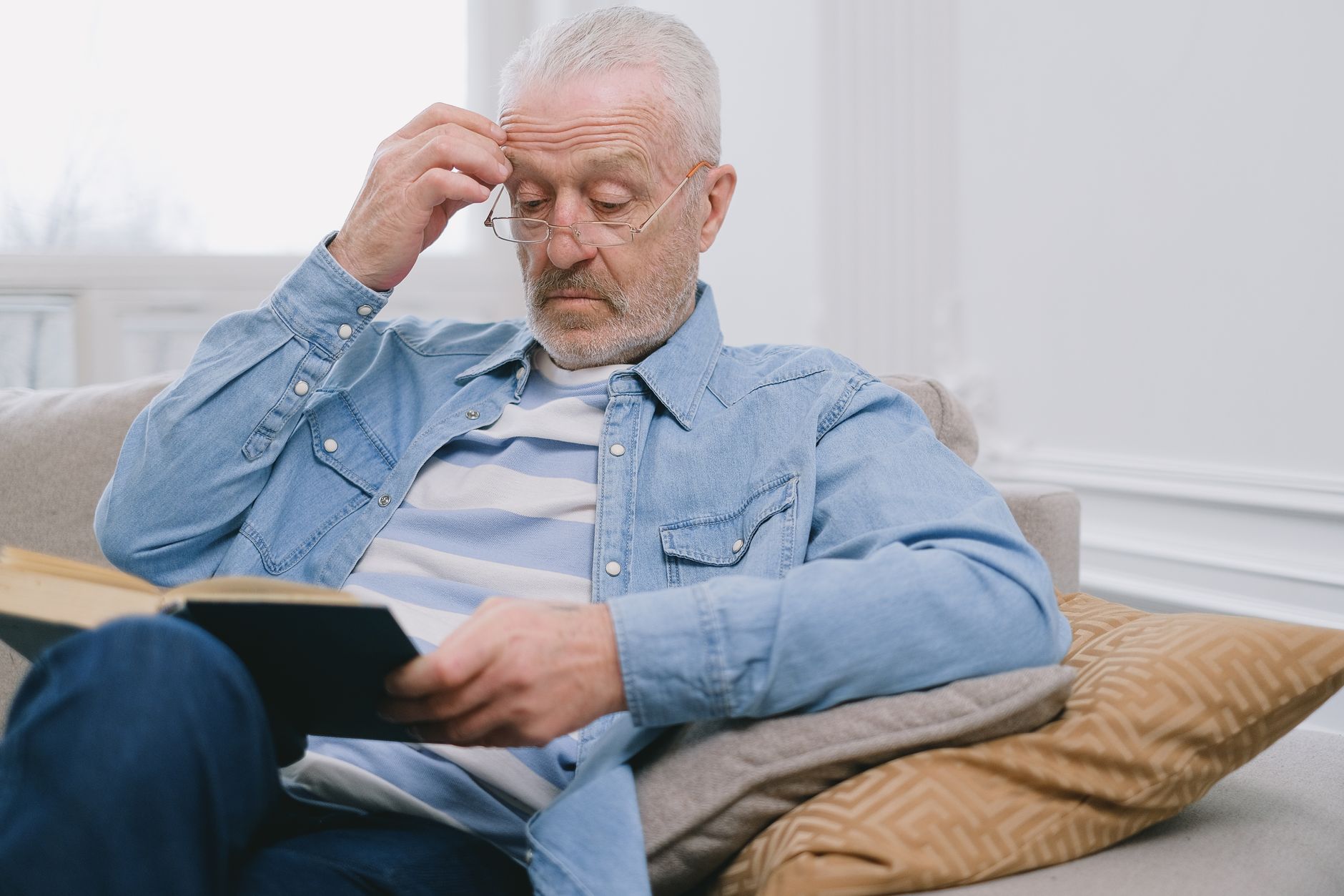 Senior man with glasses reading a book inside a stylish living room.
