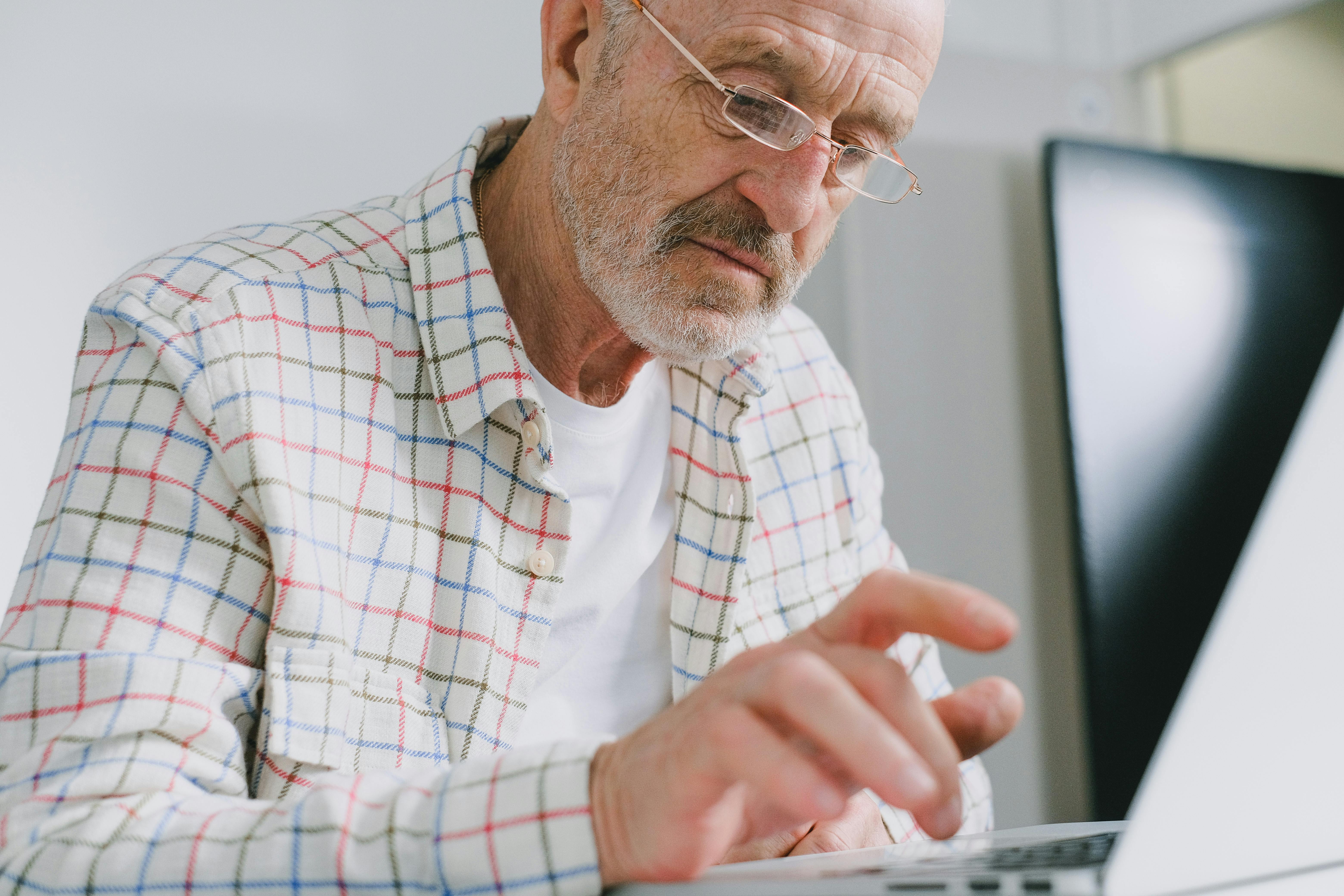 Elderly man in plaid shirt using a laptop indoors.