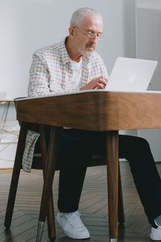 Elderly man focused on typing on laptop indoors. Natural lighting, modern ambiance.