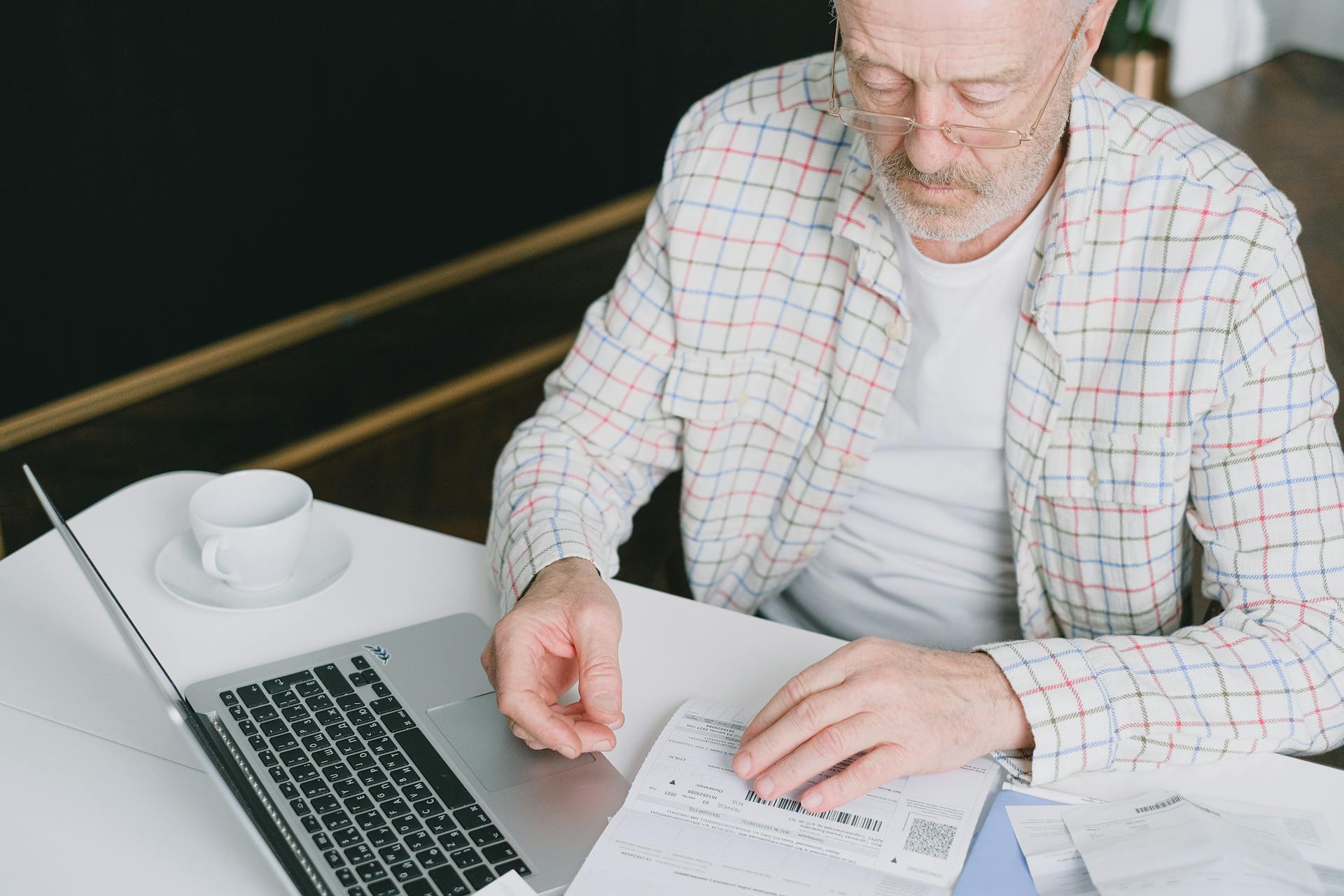 Elderly man with eyeglasses reviewing documents at a laptop. Indoor setting with natural light.