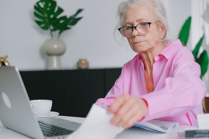 Elderly woman in pink shirt working on a laptop at home, focused and engaging with documents.