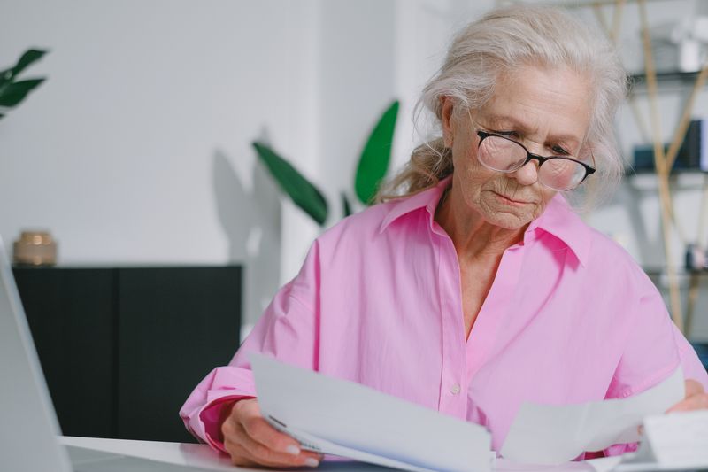 Elderly woman with eyeglasses reading papers at a desk, focusing indoors.