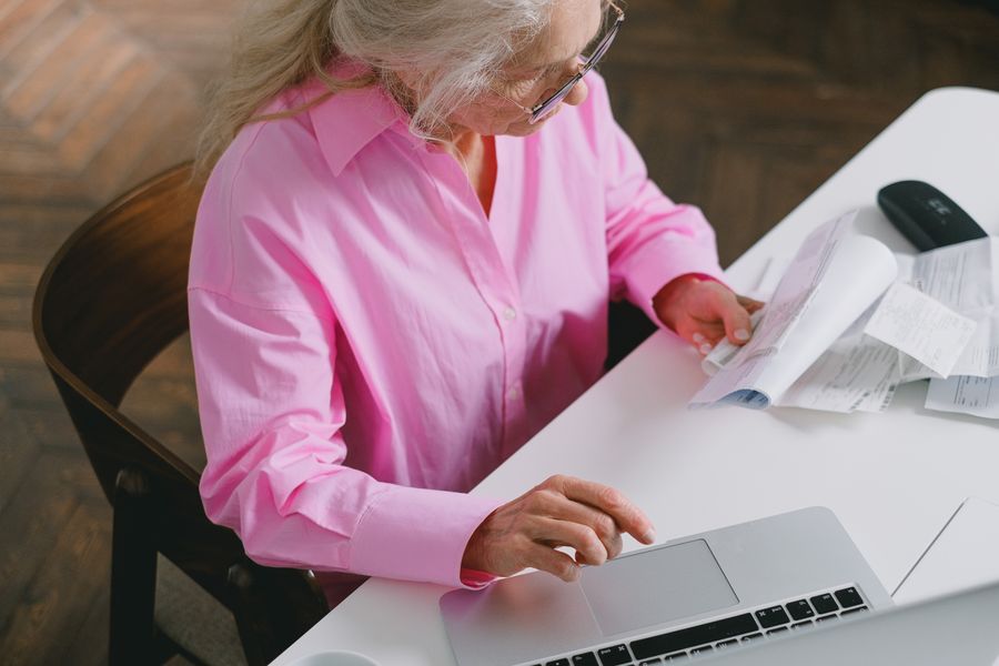 Senior woman in pink shirt using a laptop and reviewing documents at home office.