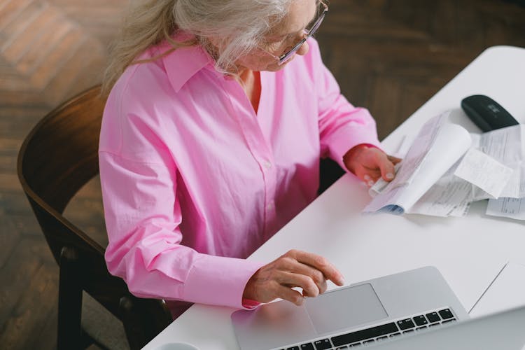 High-Angle Shot Of An Elderly Woman In Pink Long Sleeves Looking At Documents While Using A Laptop