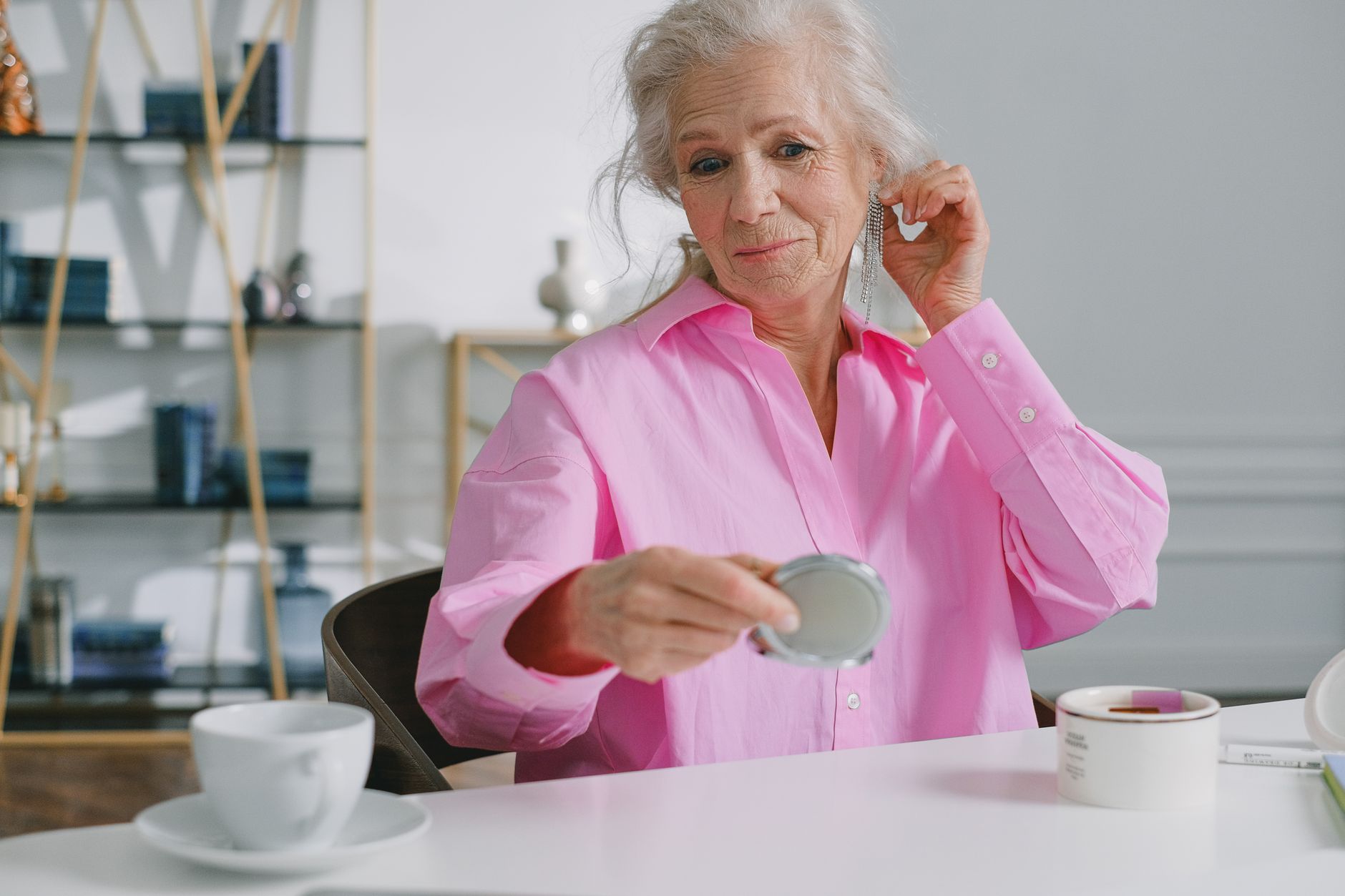A senior woman in a pink shirt using a compact mirror to admire her earrings indoors.