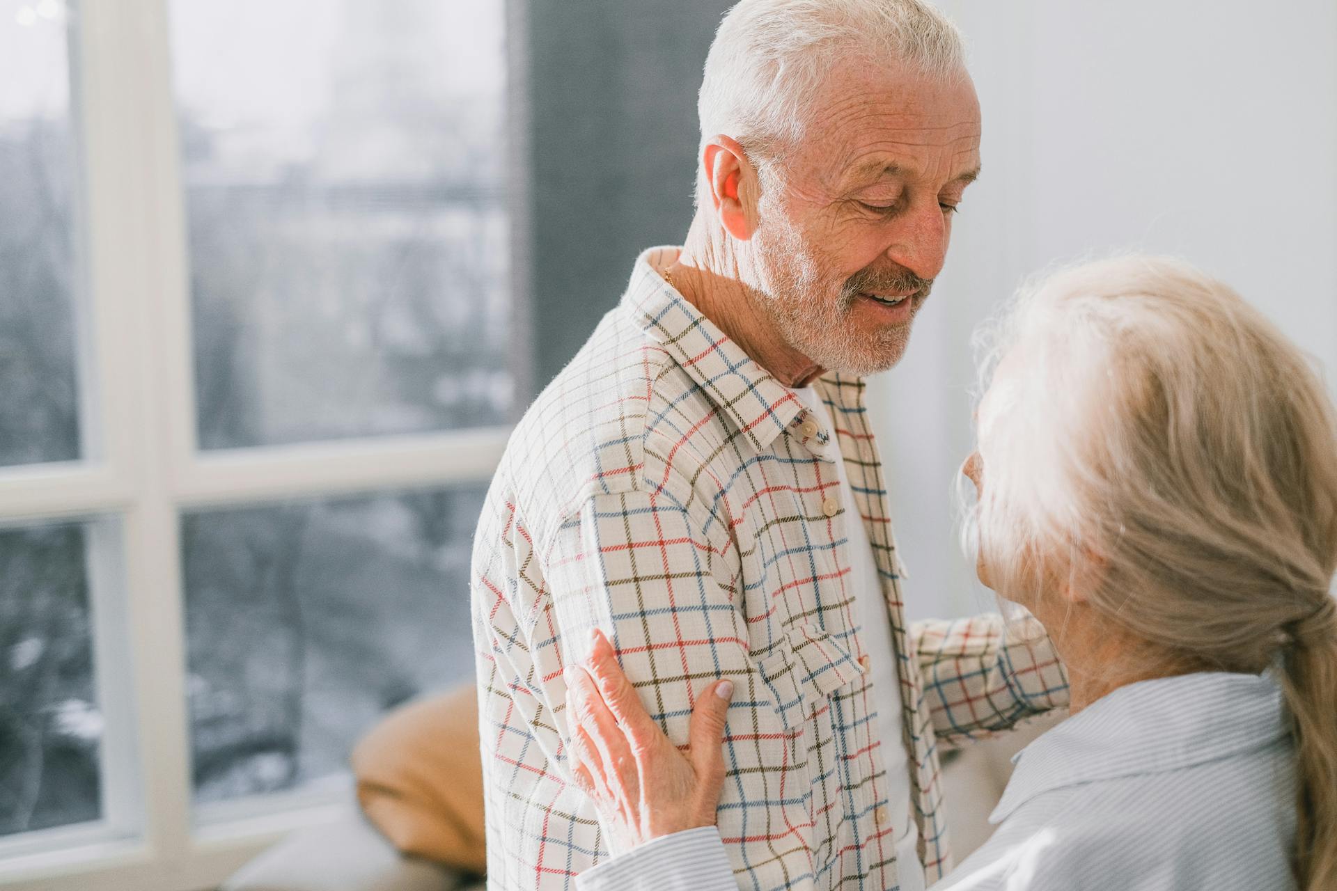 An elderly couple shares a warm embrace by a sunlit window indoors.