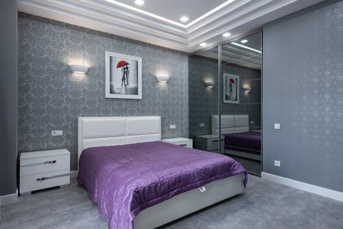 Bed with purple coverlet placed near gray wall in modern bedroom with light furniture and wardrobe with glass doors