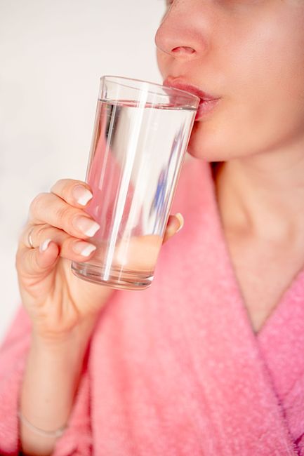 Crop female in bathrobe drinking water from glass before breakfast for heath and wellness in morning