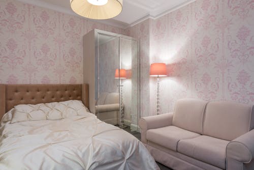 Pink floor lamp placed near soft sofa and wall in stylish bedroom with large comfortable bed with light linen