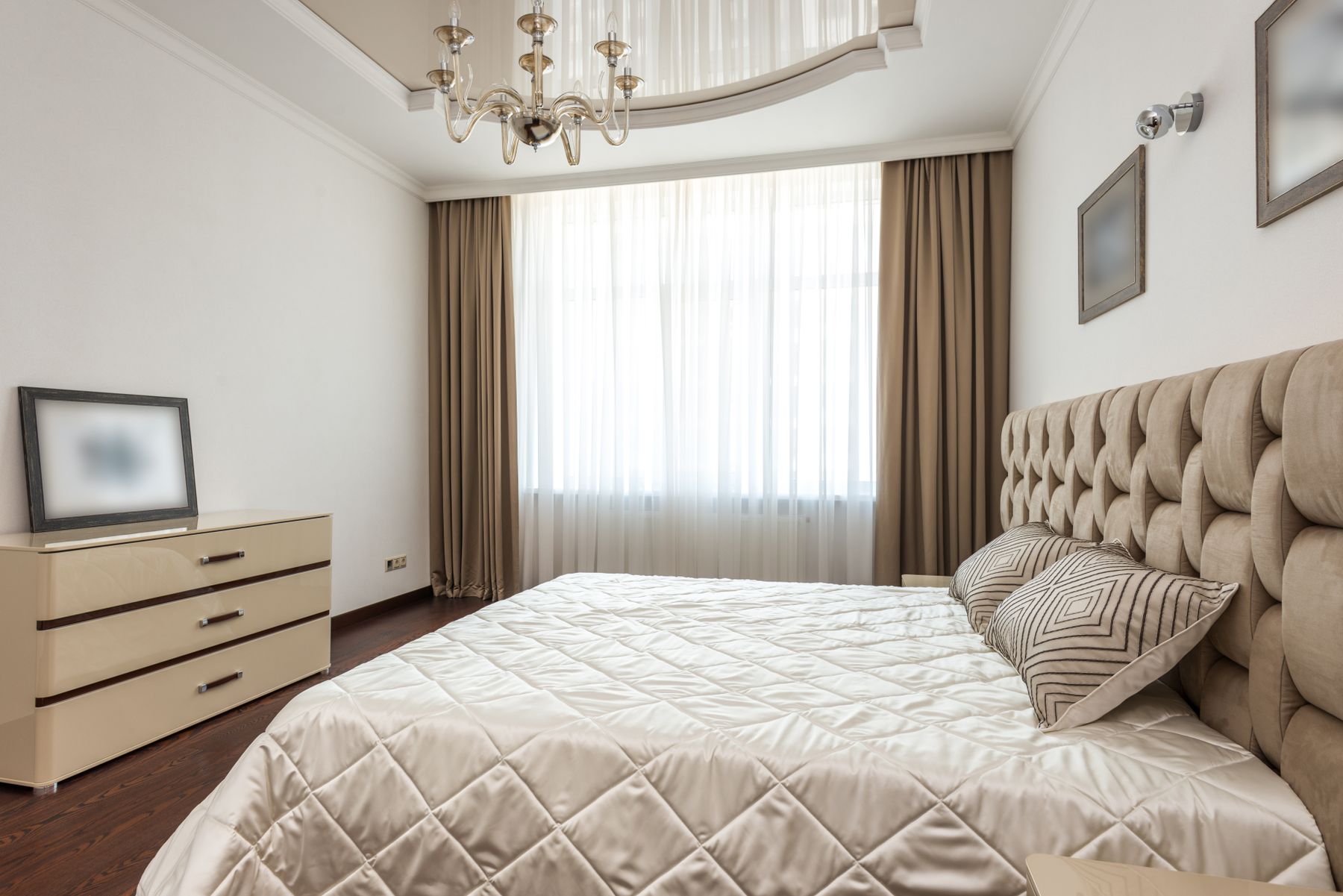 Bright spacious bedroom with a tufted beige bed, wide window, layered curtains and chandelier