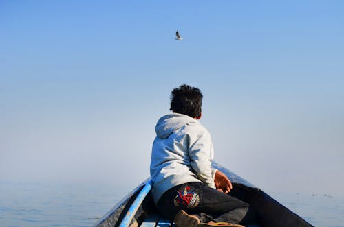Homem De Capuz Cinza E Calça Preta Sentado No Meio De Um Barco, Olhando Um Pássaro No Céu