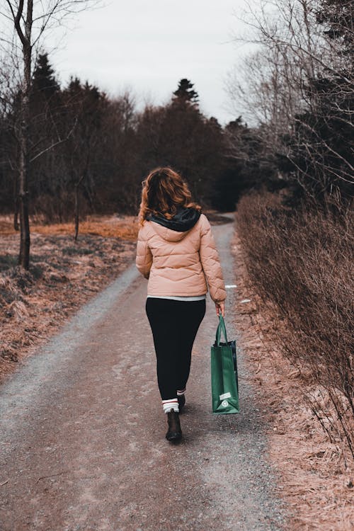 Back view full length of unrecognizable female in warm clothes and bag strolling on pathway near leafless trees and plants in autumn day in countryside