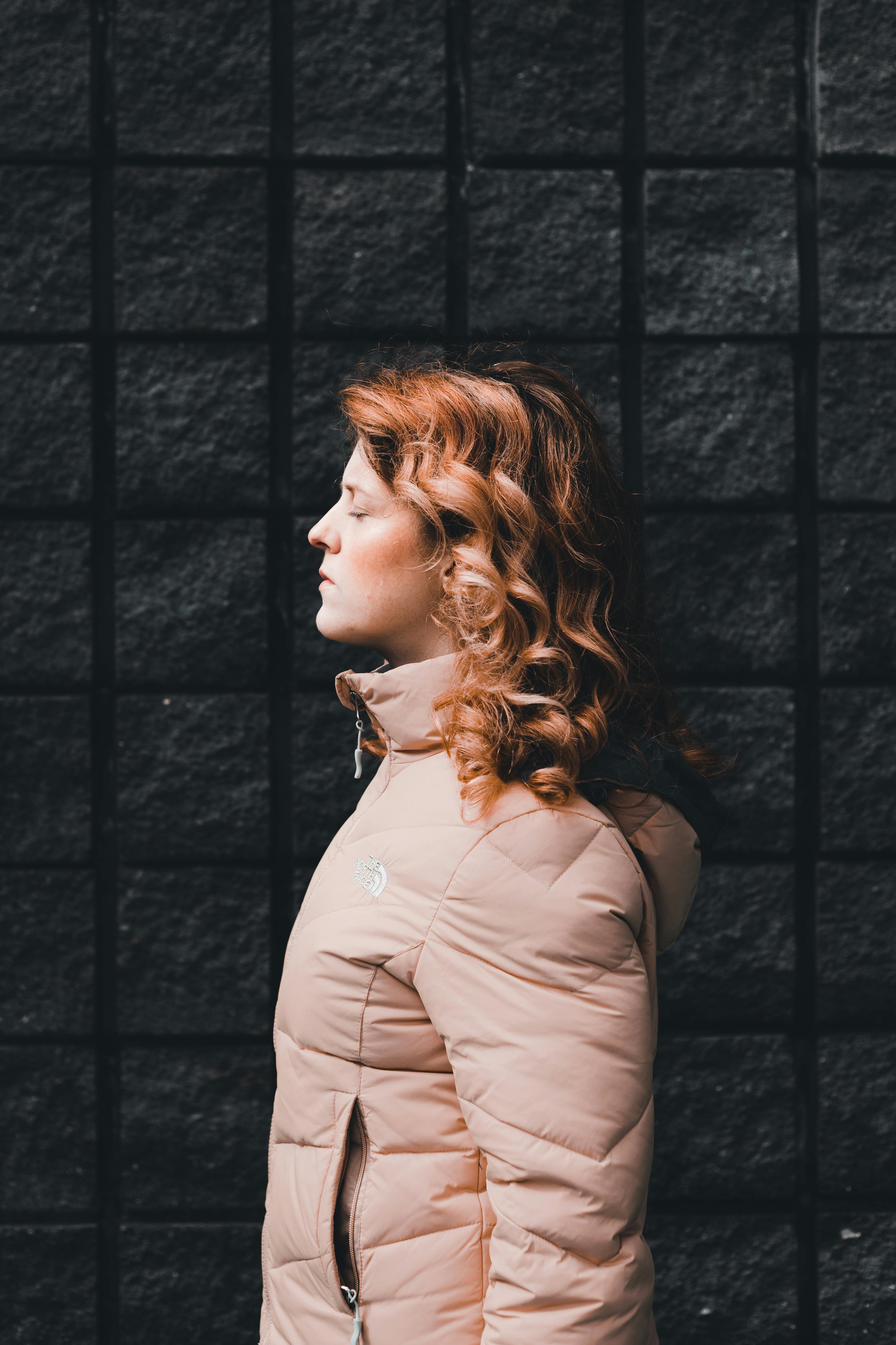 Side profile of a young woman with closed eyes wearing a pink jacket against a dark brick wall.