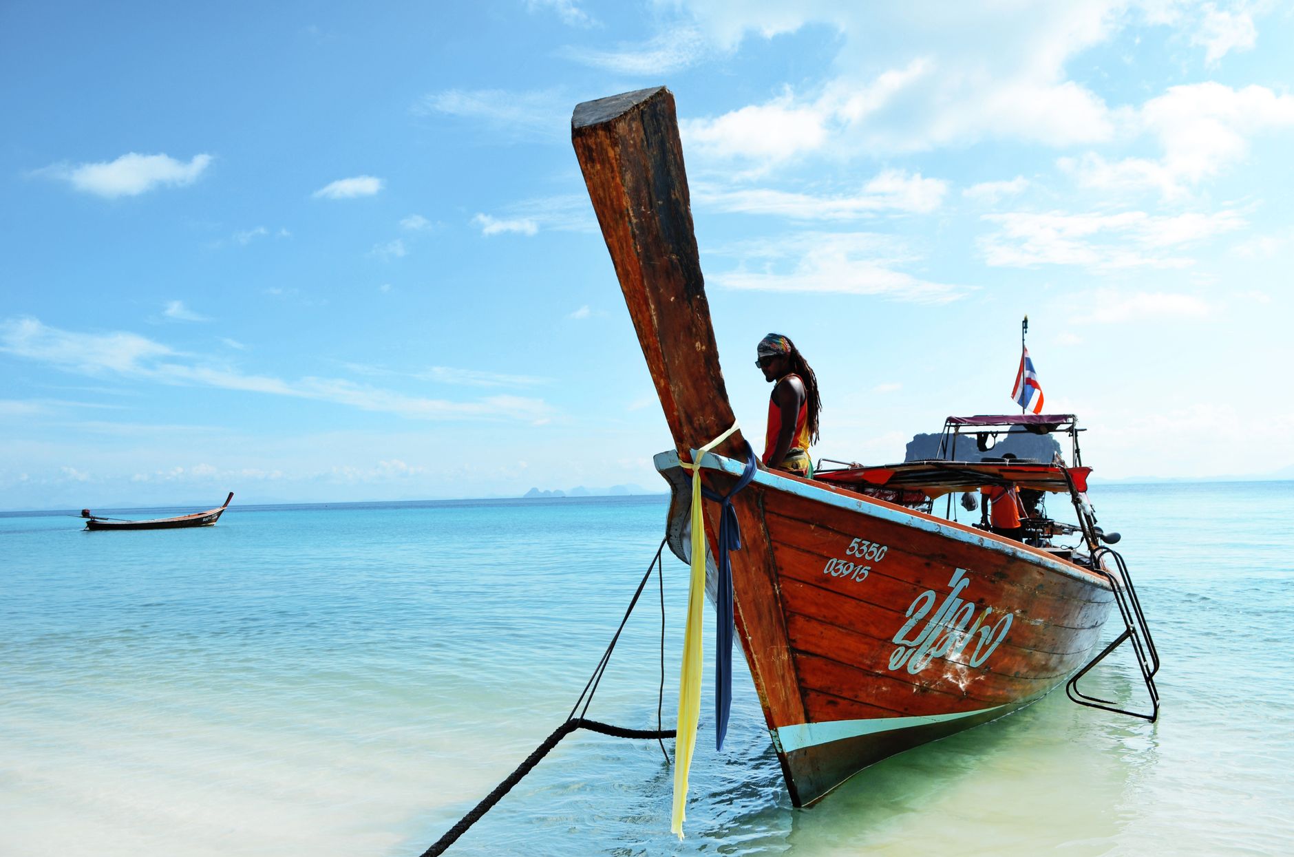 A traditional longtail boat anchored on a sunny beach in Trang, Thailand with a clear sky background.