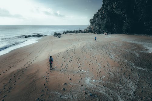 Drone view of unrecognizable travelers walking along sandy beach and admiring wavy ocean near cliff covered with lush green trees