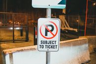 Prohibition parking road sign with Subject To Ticket inscription placed on street of city near concrete barrier in evening time