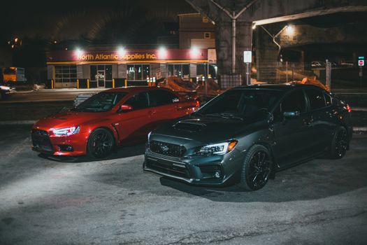 Two sport sedans, Mitsubishi Lancer EVO and Subaru WRX, parked under street lights at night.