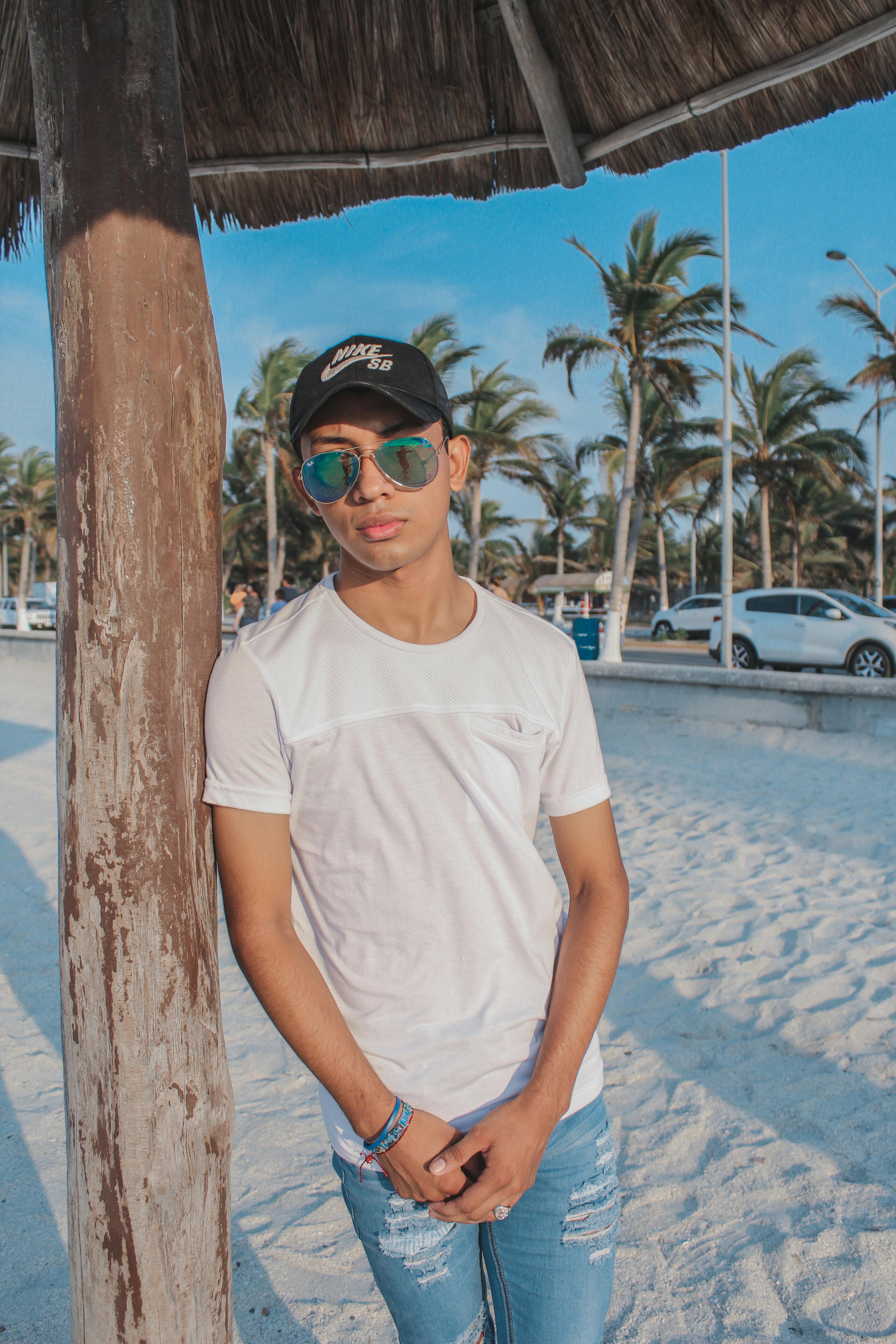 A young man in sunglasses and a white shirt stands by a palm tree on a sunny beach.