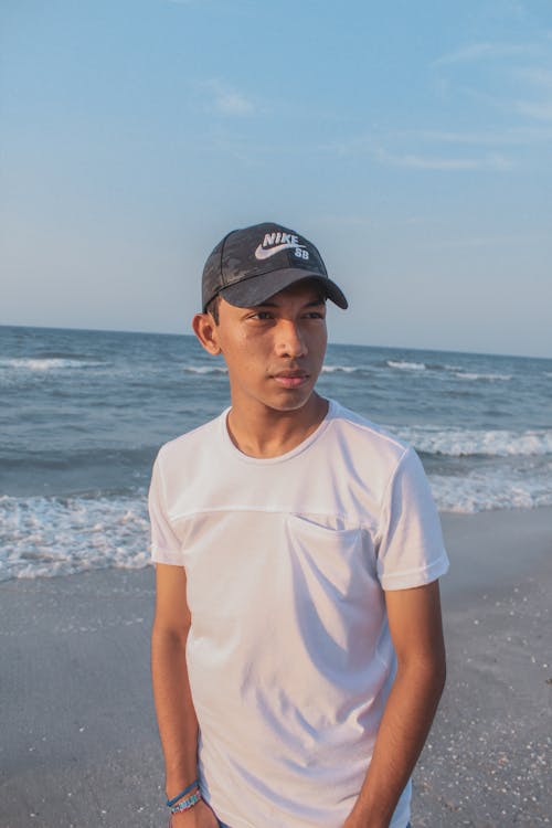 A Man in White Shirt and Black Nike Cap Standing at the Beach