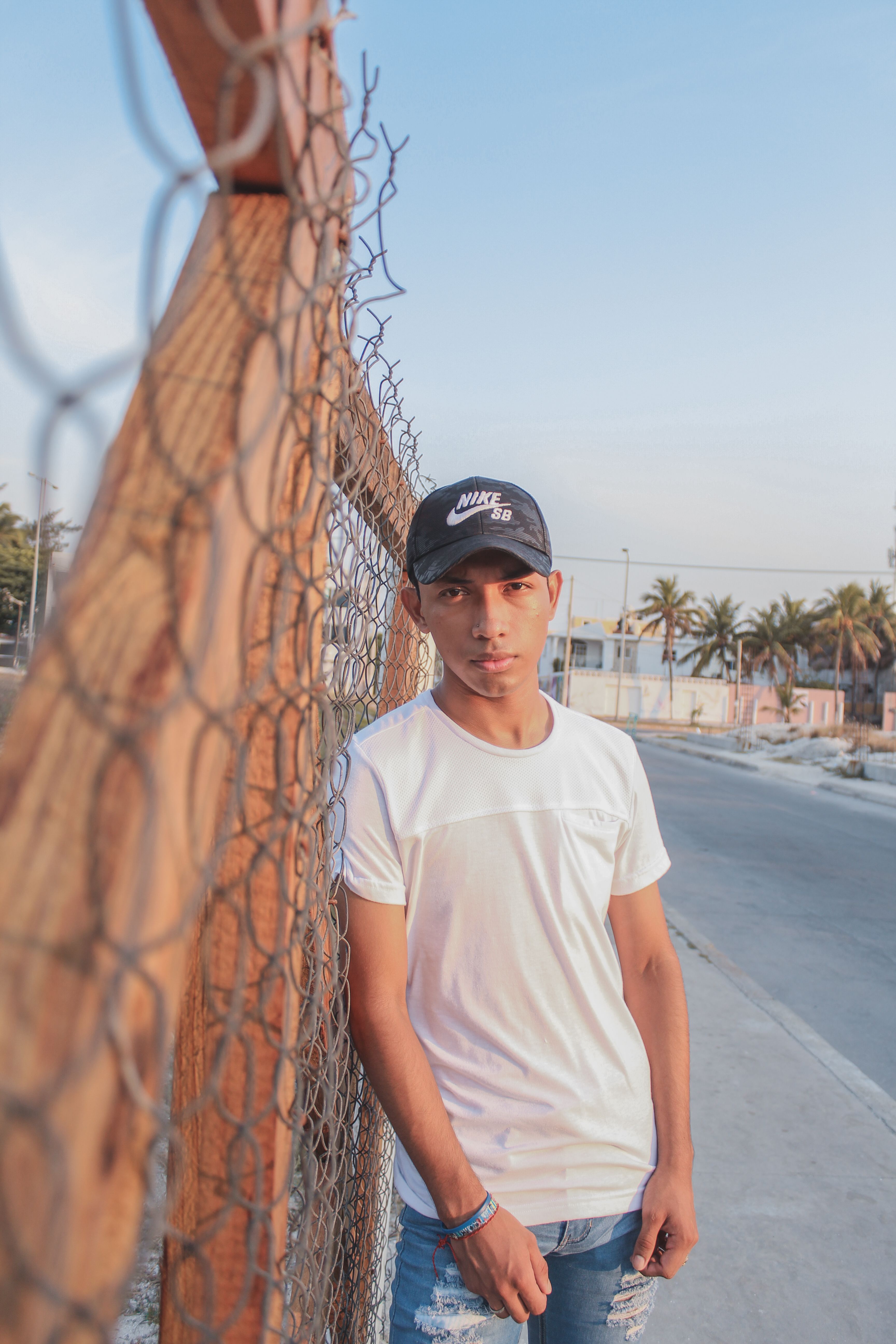 Casual young man in a cap posing by a fence outdoors during the day.