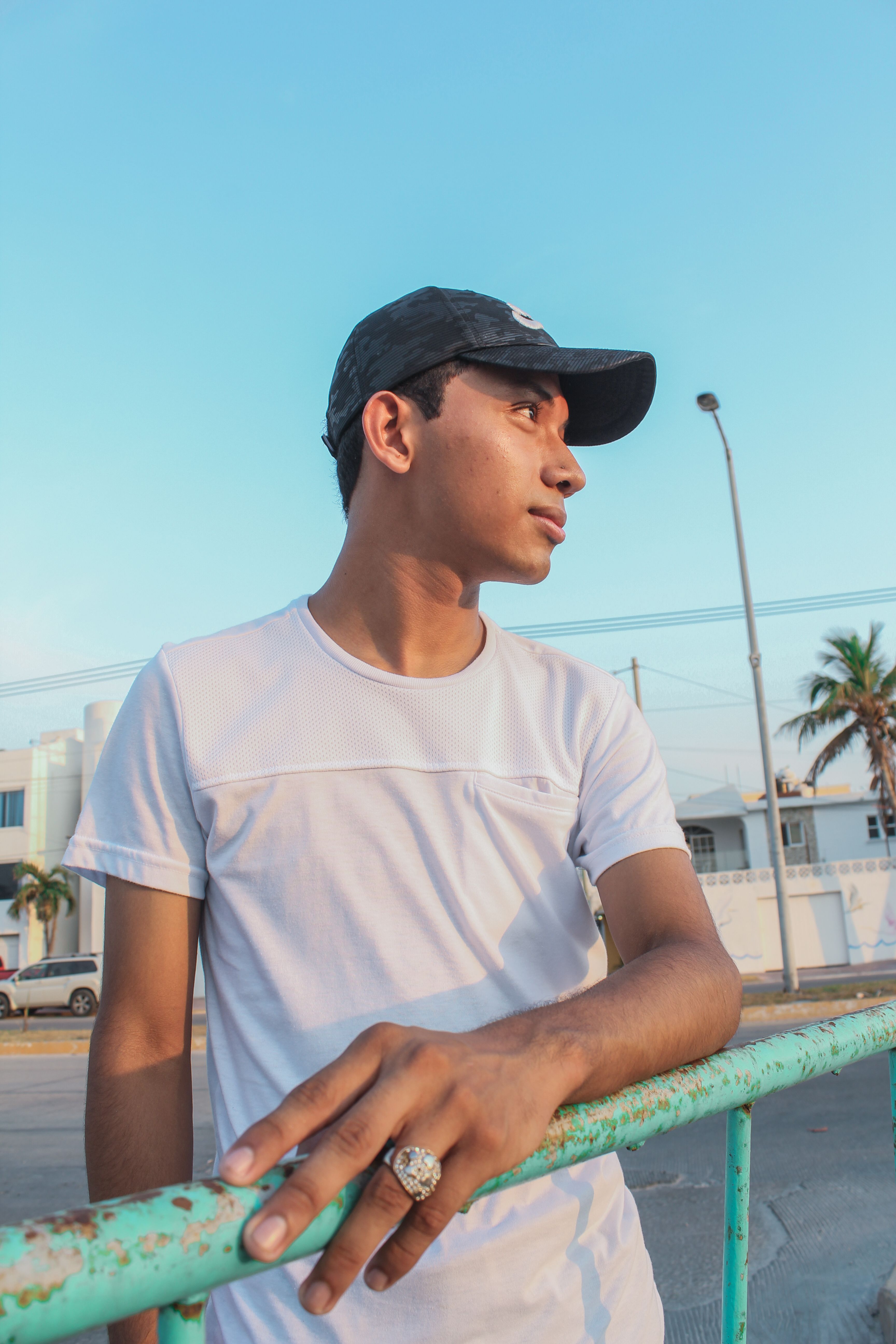 A young man in a white shirt and black cap leans thoughtfully on a railing outside.