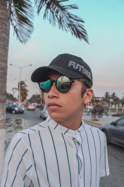 Free A young man wearing a cap and sunglasses stands outdoors in a stylish outfit, reflecting street scenery. Stock Photo