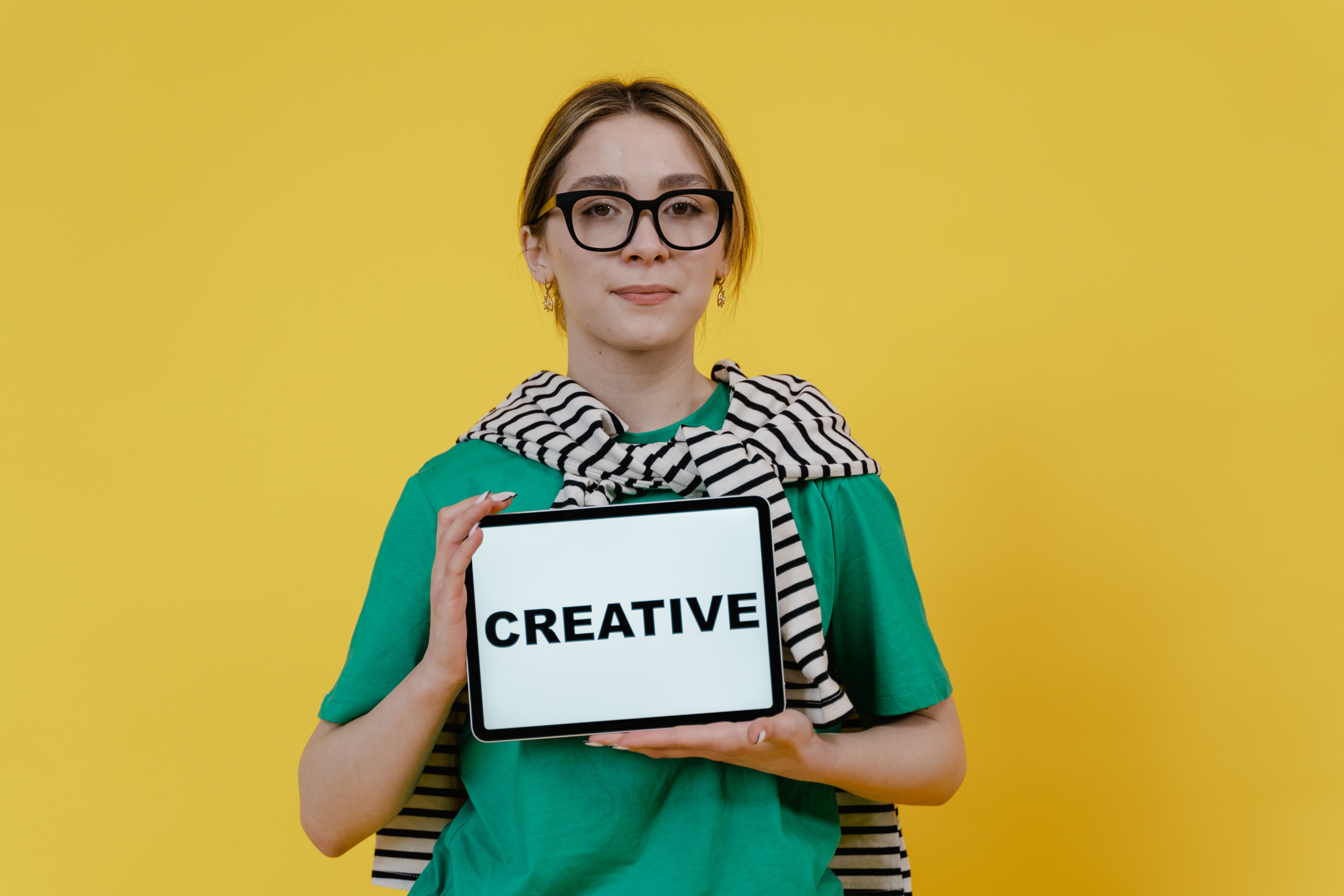 Stylish young woman in glasses holding a tablet displaying the word 'Creative' against a yellow background.
