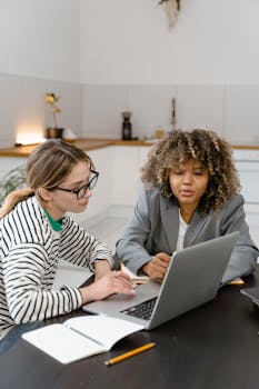 Two women discuss work strategy at a laptop in a modern office.