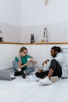 Two colleagues having a casual meeting on a kitchen floor, discussing documents and using a laptop.