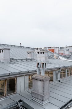 Detailed view of city rooftops with metal chimneys and windows, under cloudy sky.