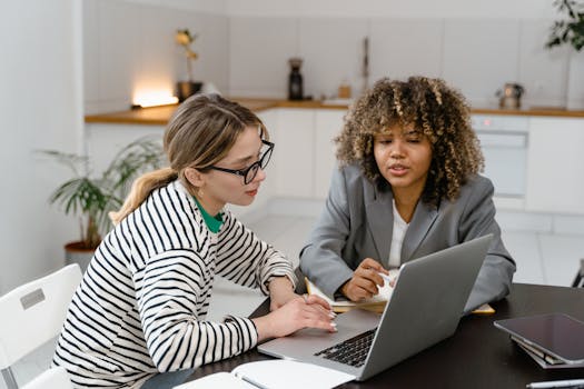 Two women engaged in a workplace discussion, working on a laptop together indoors.