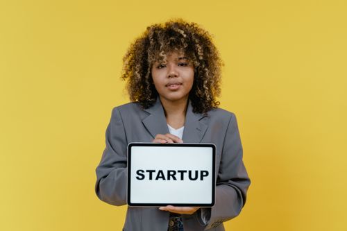 Free Portrait of a woman holding a tablet showing the word 'STARTUP' against a yellow background. Stock Photo