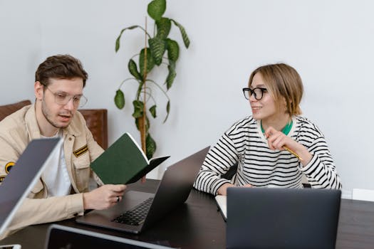 Two young adults collaborating with laptops and notebooks in an office space.