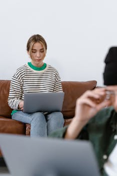 Young woman sitting on a couch focused on her laptop, working remotely with a second person blurred in foreground.