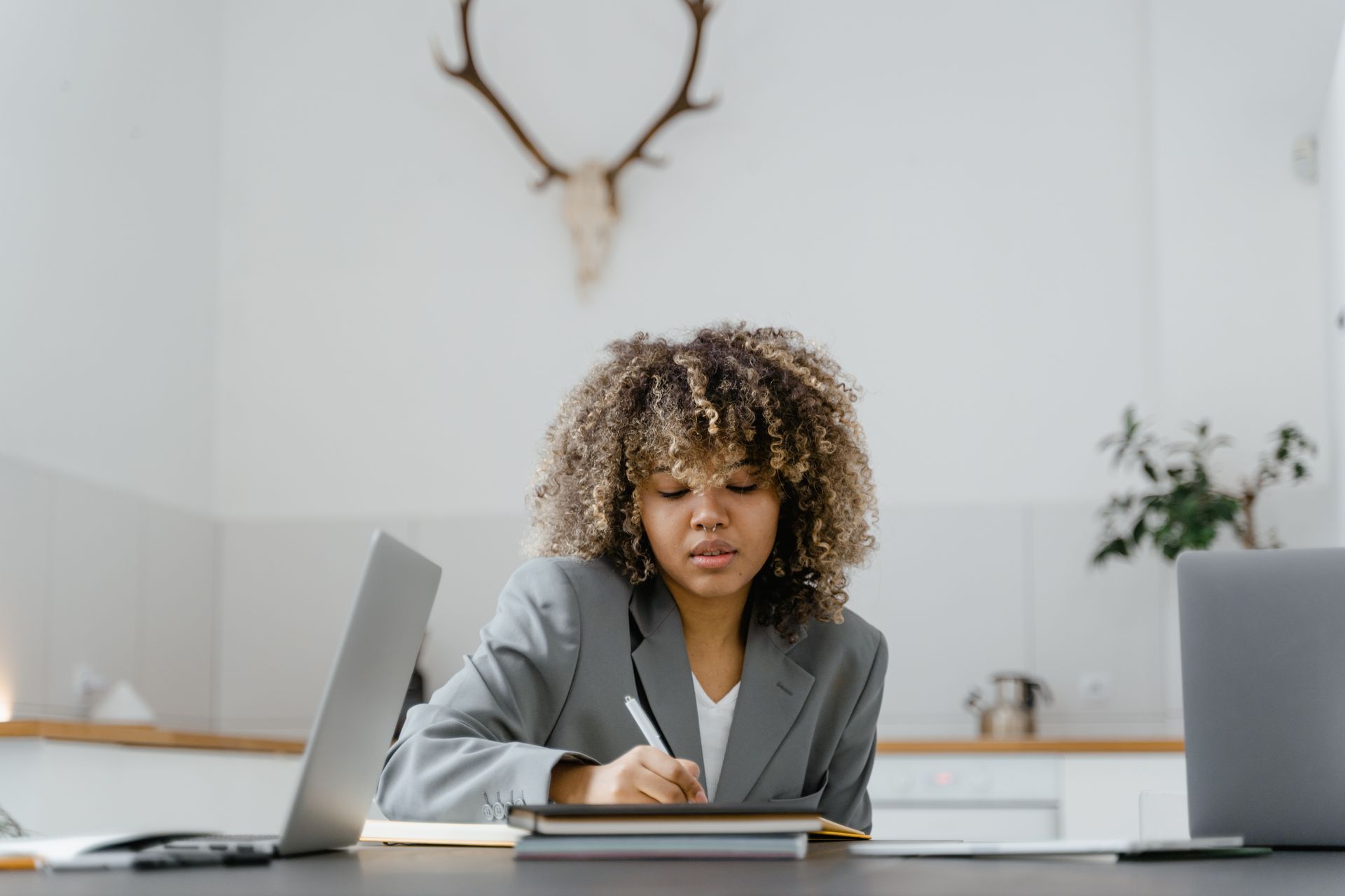 A woman with curly hair writing notes at a desk with laptops in a stylish office setting.