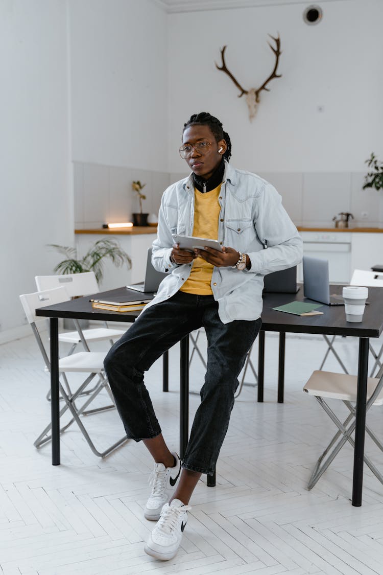 A Man In Denim Jacket And Black Pants Sitting On The Table While Holding A Tablet